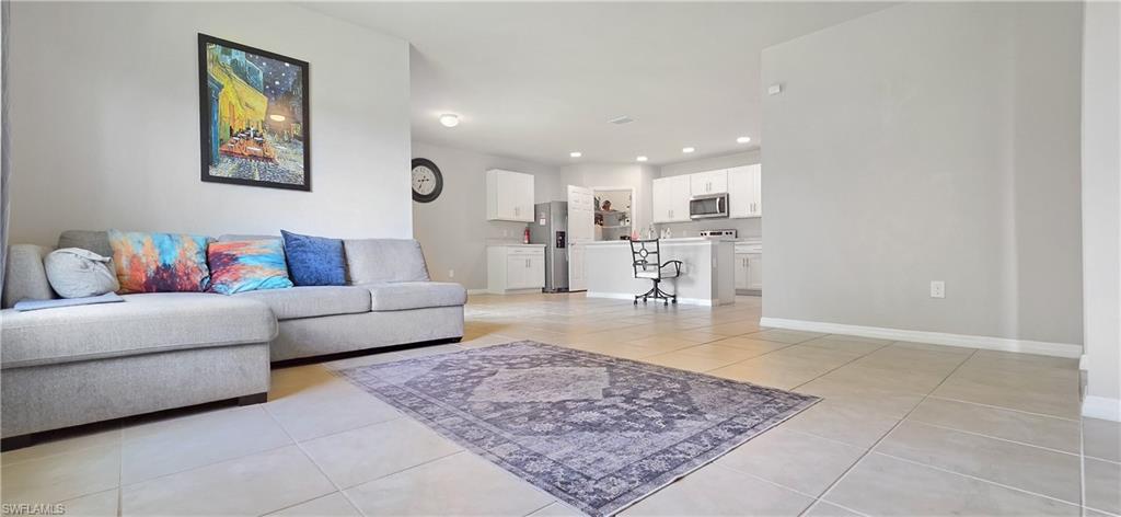 Open-concept living space featuring neutral tile flooring throughout, a sectional sofa with decorative pillows, area rug, and a well-appointed kitchen with stainless steel appliances, white cabinetry, and pendant lighting