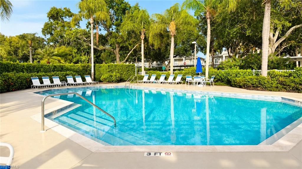 Resort-style community pool featuring crystal-clear waters, spacious sun deck with lounge seating, mature tropical landscaping including palm trees, and lush perimeter hedging for privacy