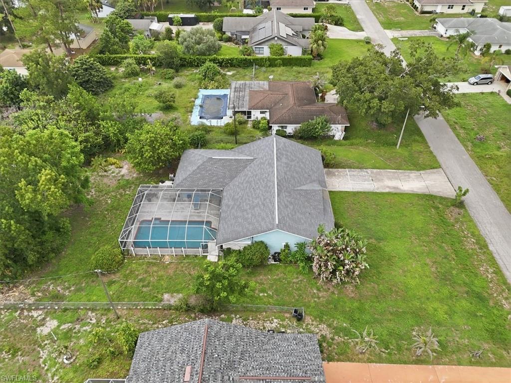 Aerial view showcasing this residential property featuring a gray composite roof, enclosed pool with retractable cover, manicured lawn, mature landscaping with established trees, and substantial lot size with single-story construction