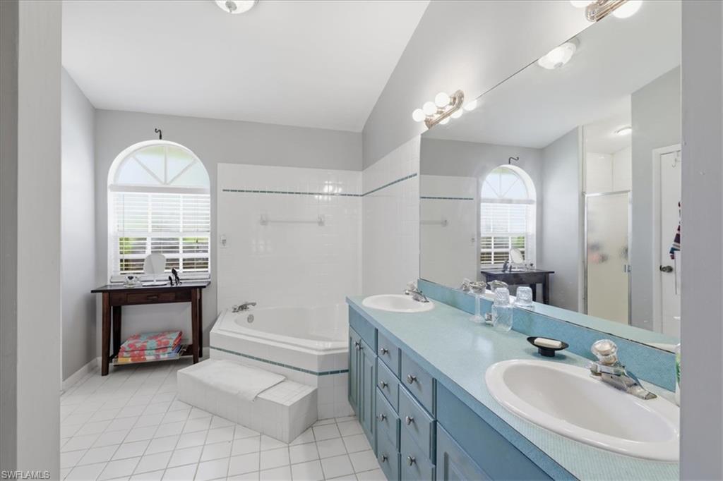 Bathroom featuring a dual vanity with a blue countertop and integrated sinks, a built-in corner tub with tiled steps, white tiled flooring, and an arched window with horizontal blinds