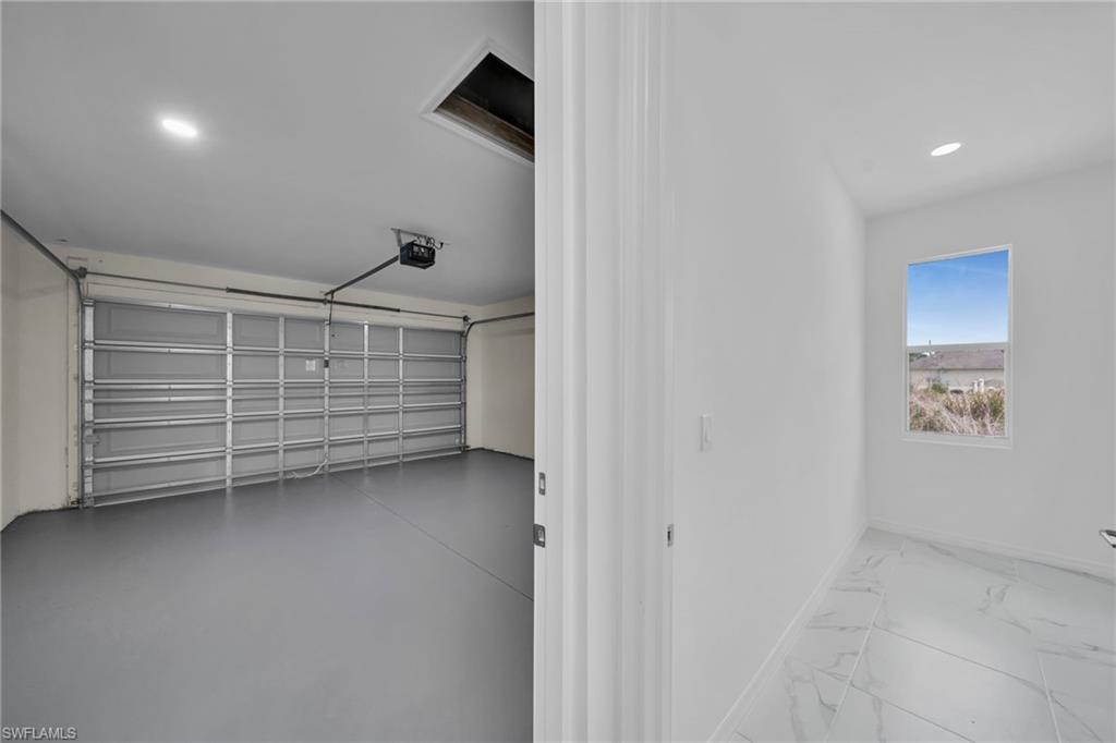 Spacious two-car garage with aluminum sectional door and epoxy-coated flooring, adjoining bright hallway featuring polished stone-finish flooring, recessed lighting, and large window framing desert landscape views