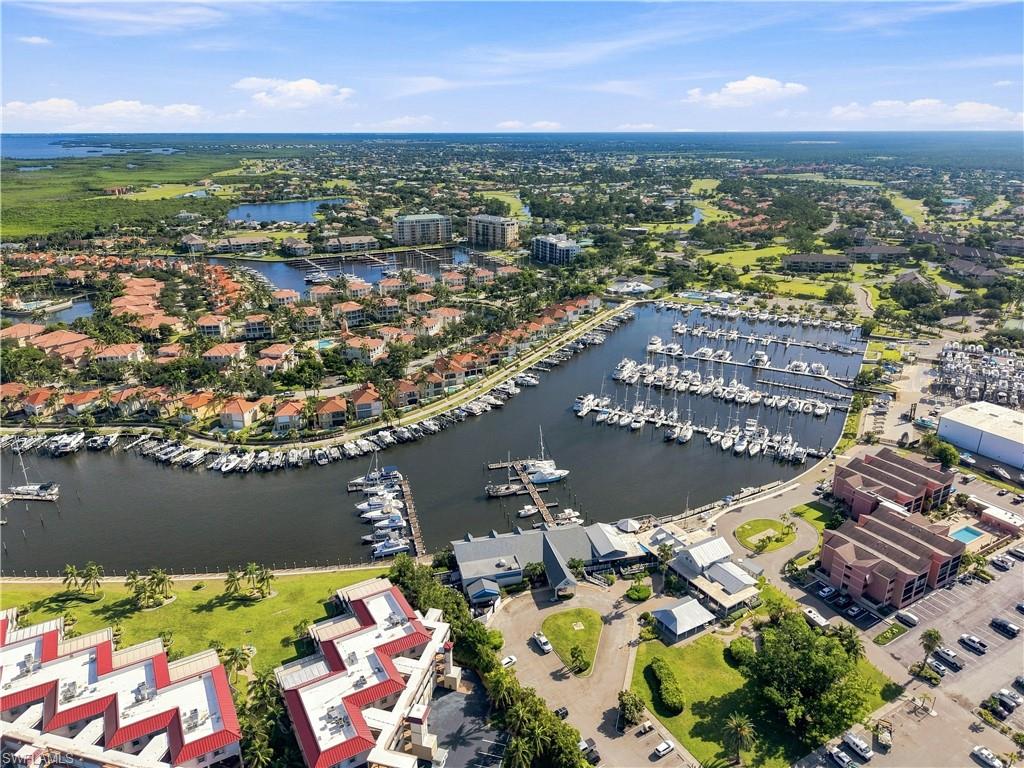 Aerial perspective featuring a marina with numerous boats, waterfront residences with red tile roofs, expansive green spaces, and coastal water bodies in the distance
