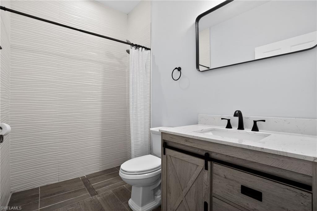 Wavy white tile shower surround, wood-finish vanity with sliding barn-door style cabinet, black framed rectangular mirror, black hardware and fixtures, and dark wood-look tile flooring