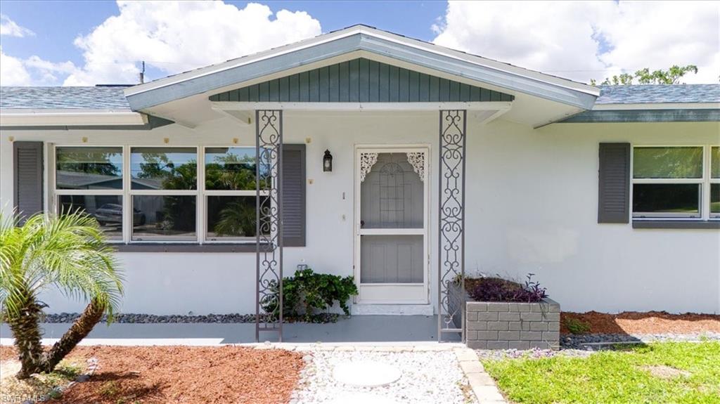 White stucco exterior with a gray trim, featuring a screened entry, decorative metal columns, and a front yard with mixed landscaping