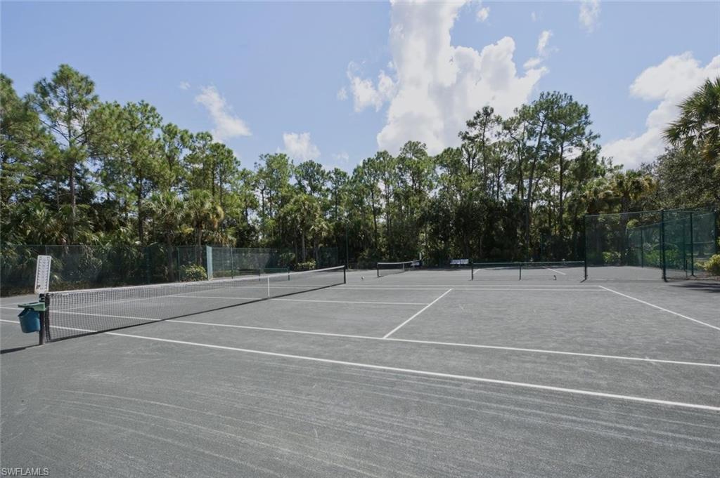 Clay tennis court with white line markings, surrounded by green fencing and mature trees