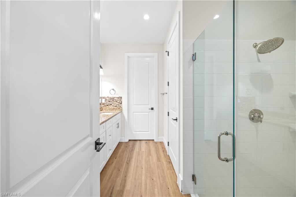 Wood-finish flooring, white flat-panel cabinetry, a light-toned countertop, a glass-enclosed shower, and a rainfall showerhead