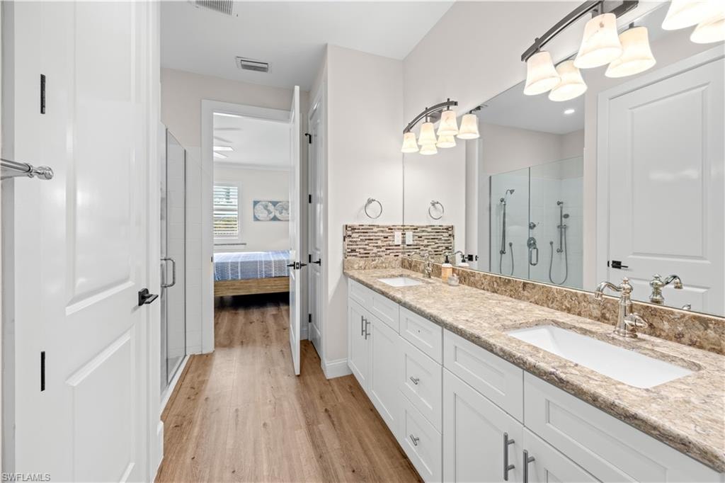 Dual vanity with integrated sinks, wood-finish flooring, glass-enclosed shower, stone-finish countertop, and decorative tile backsplash