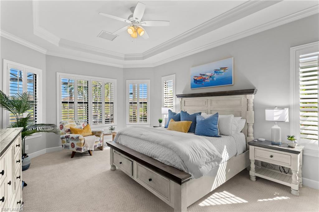 Tray ceiling with crown molding, ceiling fan, multiple windows with plantation shutters, carpeted flooring, and a built-in dresser