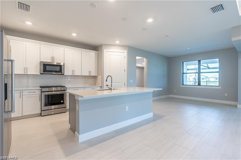 Open-concept kitchen featuring white shaker cabinetry, stainless steel appliances, a center island with a sink, and light-toned tile flooring