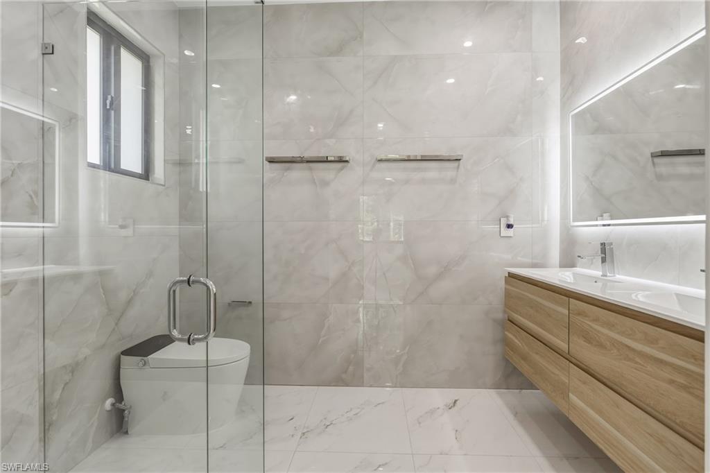 Glass-enclosed shower, floating wood-finish vanity, integrated sink, illuminated mirror, and marble-look tile flooring