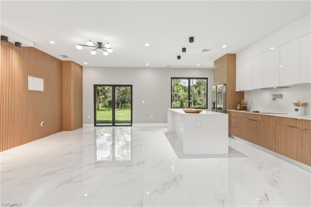 Expansive open-concept living area featuring white marble-look flooring, a modern kitchen island with an integrated sink, dual-tone cabinetry, and a full-light glass door