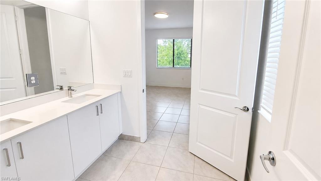 Dual vanity with white cabinetry and integrated sinks, large wall-mounted mirror, light-toned tile flooring, and a large window with blinds