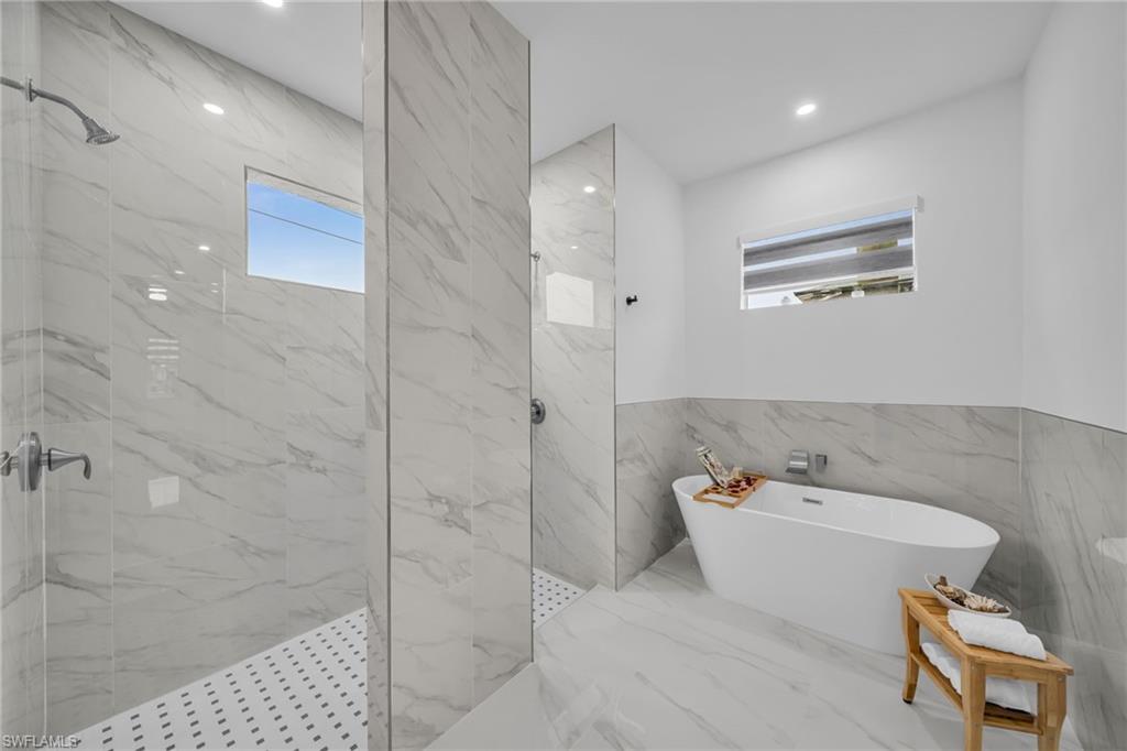 Freestanding soaking tub, walk-in shower with tiled walls, separate shower stall with glass paneling, recessed lighting, and a window with roller shades