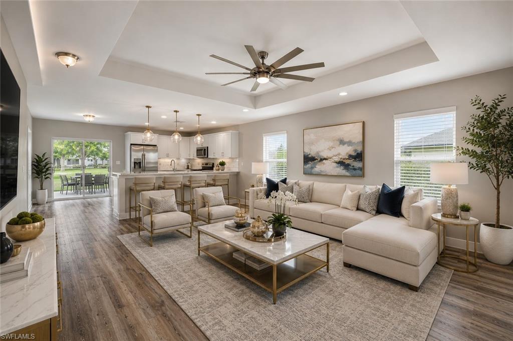 Open-concept great room featuring soaring tray ceilings, luxury vinyl plank flooring, and abundant natural light. The spacious living area flows seamlessly into the designer kitchen with quartz countertops, white shaker cabinetry, stainless steel appliances, a large breakfast bar with seating, and modern pendant lighting. Sliding glass doors open to the covered lanai. [Virtually Staged]