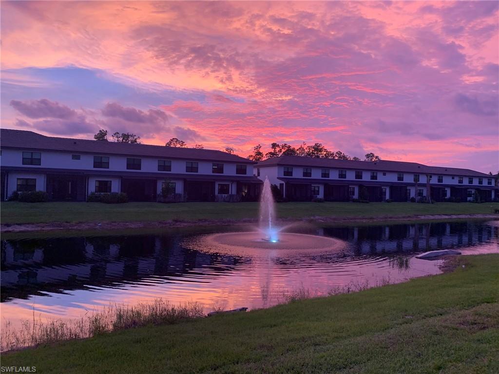 Picturesque grounds featuring manicured lawns, a tranquil pond with an illuminated fountain, and a row of residential buildings silhouetted against the evening sky