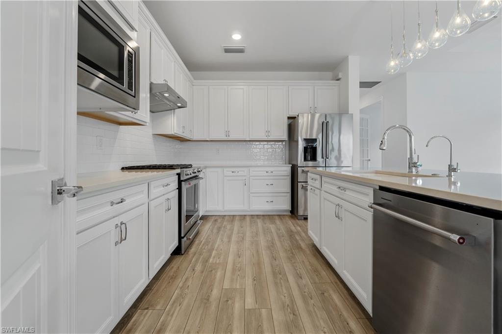 Kitchen featuring white shaker cabinetry, stainless steel appliances, wood-finish flooring, a center island with sink, and pendant lighting