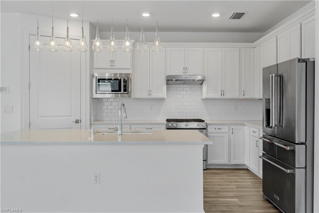 White shaker cabinetry with crown molding, stainless steel appliances, subway tile backsplash, and a large island with sink and pendant lighting