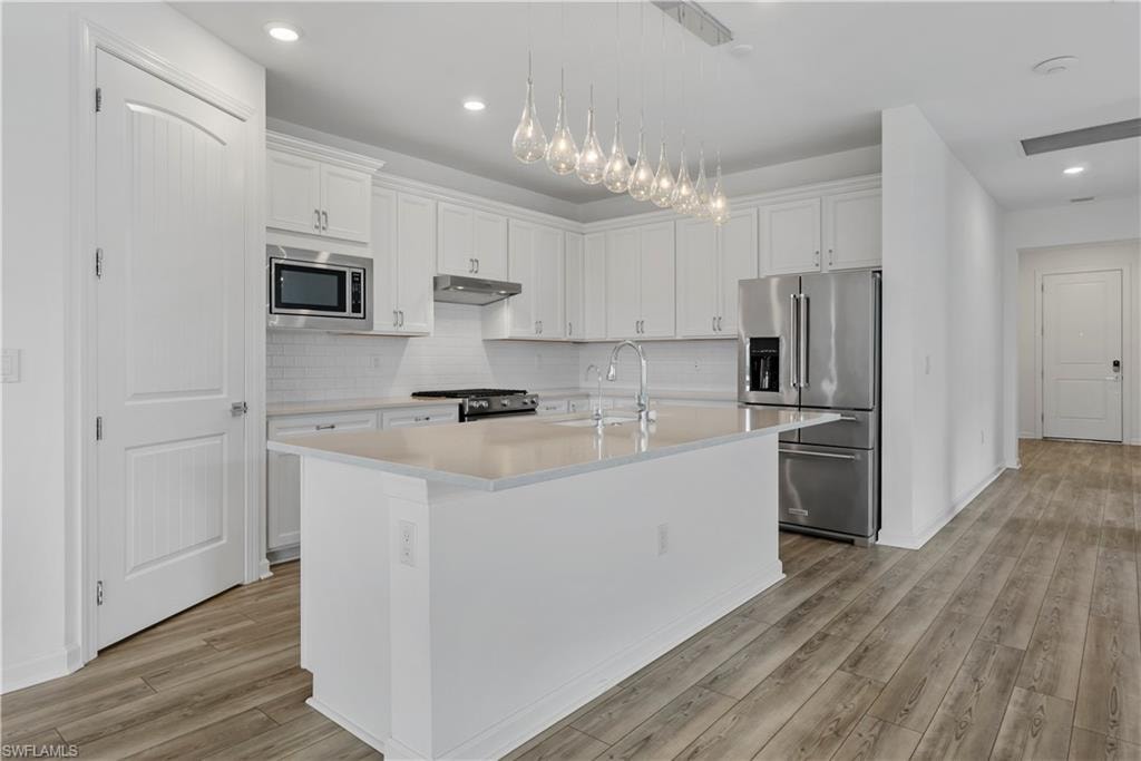 White kitchen cabinetry with stainless steel appliances, central island, pendant lighting fixture, and wood-finish flooring
