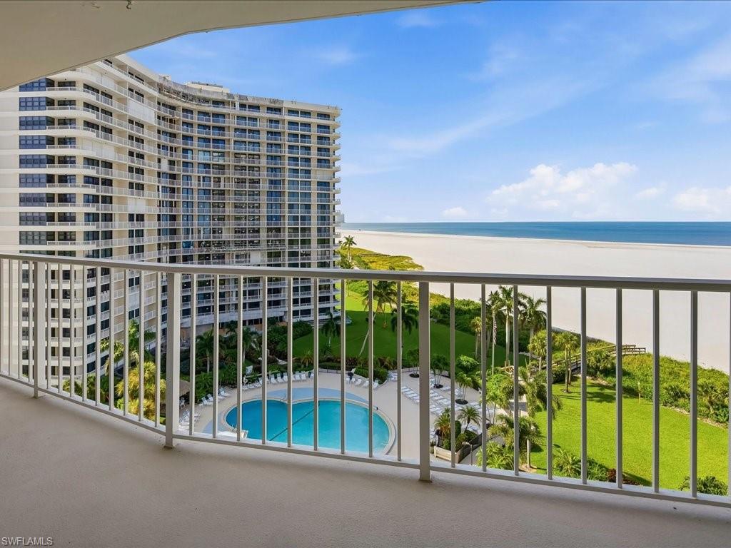 Expansive balcony with white metal railings, overlooking a large resort-style swimming pool, lush green landscaping, and a broad sandy beach with ocean beyond