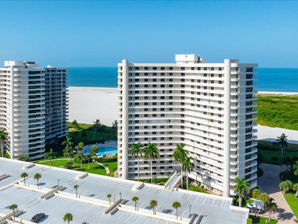 High-rise coastal building with multiple private balconies, expansive parking structure, direct beach access, and a resort-style pool with surrounding landscaping