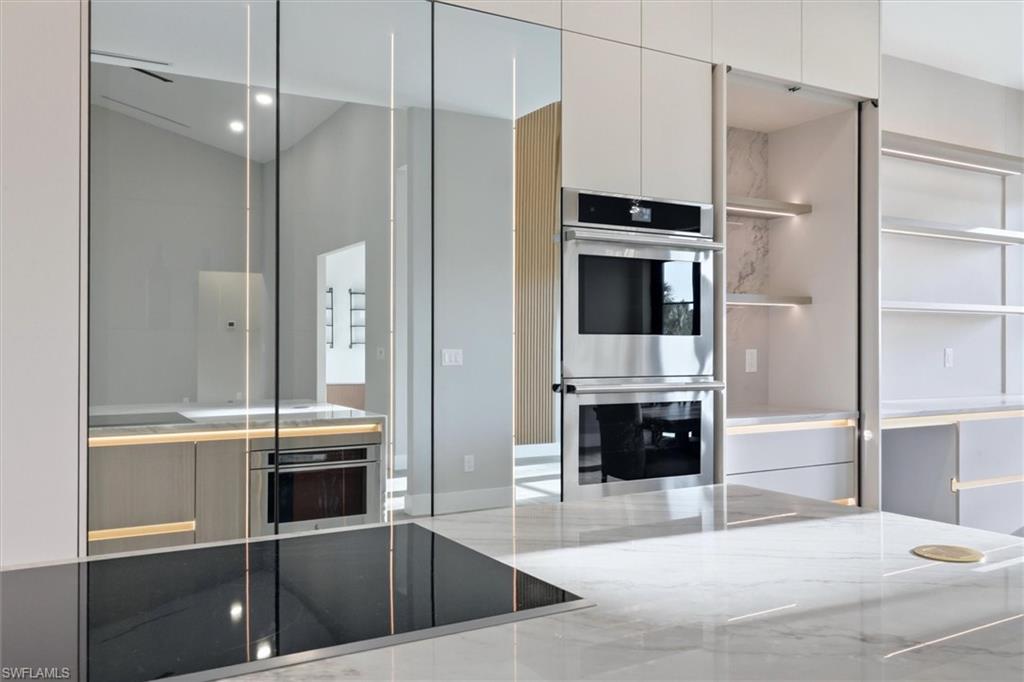 Contemporary kitchen featuring floor-to-ceiling glass cabinetry with integrated lighting, dual stacked wall-mounted ovens with stainless steel finishes, a waterfall-edge quartz countertop island, and polished concrete flooring throughout the open culinary space