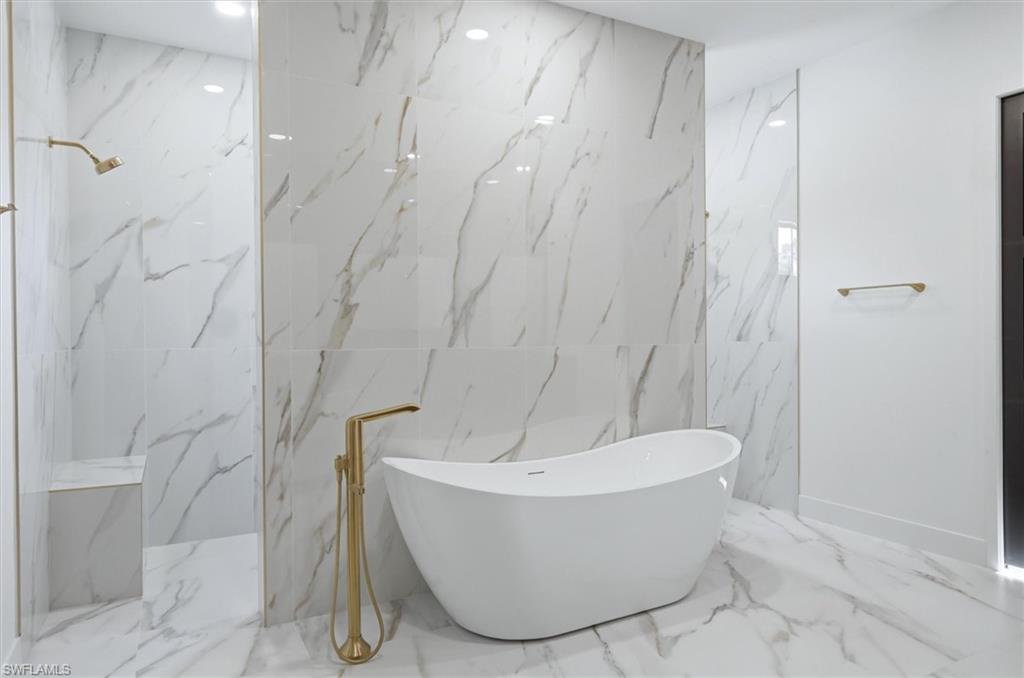 Freestanding soaking tub with a floor-mounted gold-tone faucet, walk-in shower featuring a built-in bench, and large format marble-look tile flooring and walls