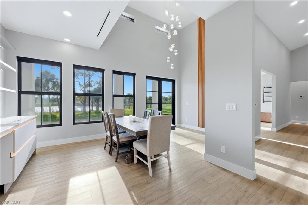 Modern open-concept dining and kitchen area featuring floor-to-ceiling black-framed windows with expansive natural light, wood-finish flooring throughout, a contemporary pendant light fixture, a vertical wood-grain accent column, and seamless sightlines to additional living spaces