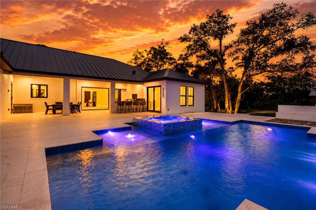Luxurious resort-style pool with vibrant LED lighting and spa features, framed by a modern architectural residence with expansive glass sliding doors, metal roof accents, and illuminated interior spaces