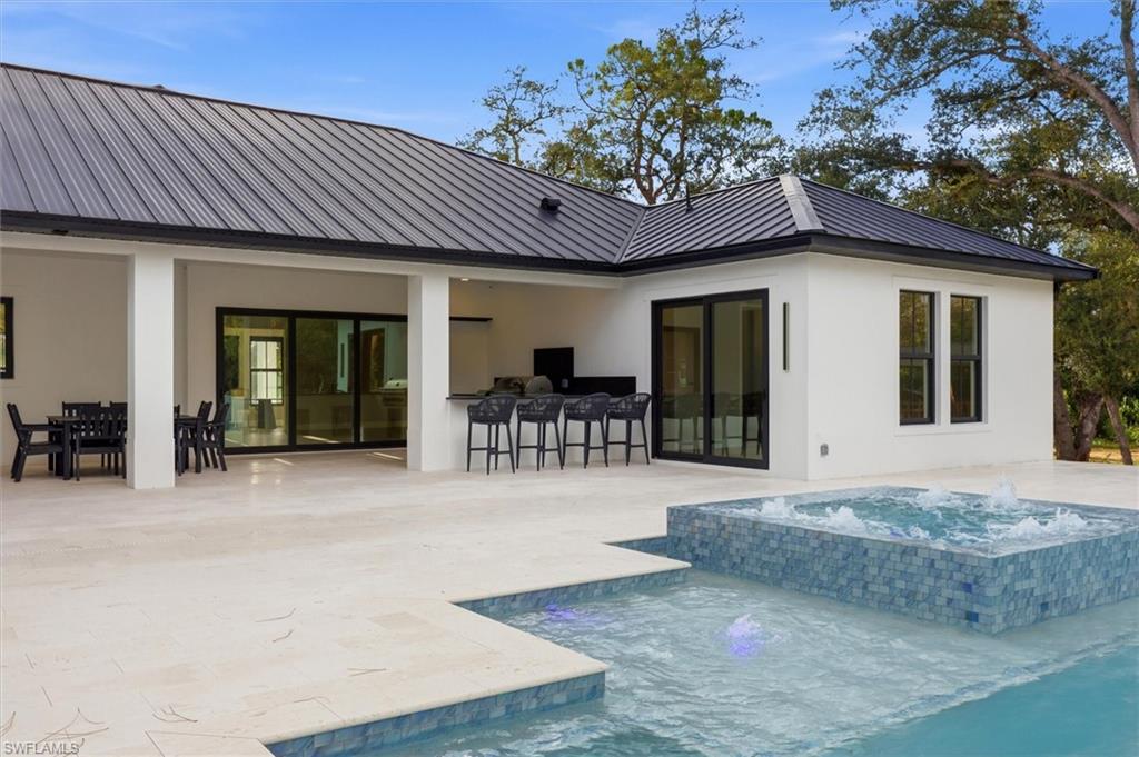 Modern home with expansive outdoor living spaces featuring a resort-style pool and spa with stone accents, wide travertine-finish patio, multiple glass sliding door systems, black metal roof, white exterior walls, and mature landscaping with native trees