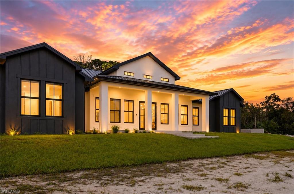 Modern farmhouse residence featuring a striking black and cream exterior with board-and-batten siding, expansive front-facing windows with illuminated frames, gabled roofline with clerestory detailing, manicured lawn, and mature landscape framing on a generous lot