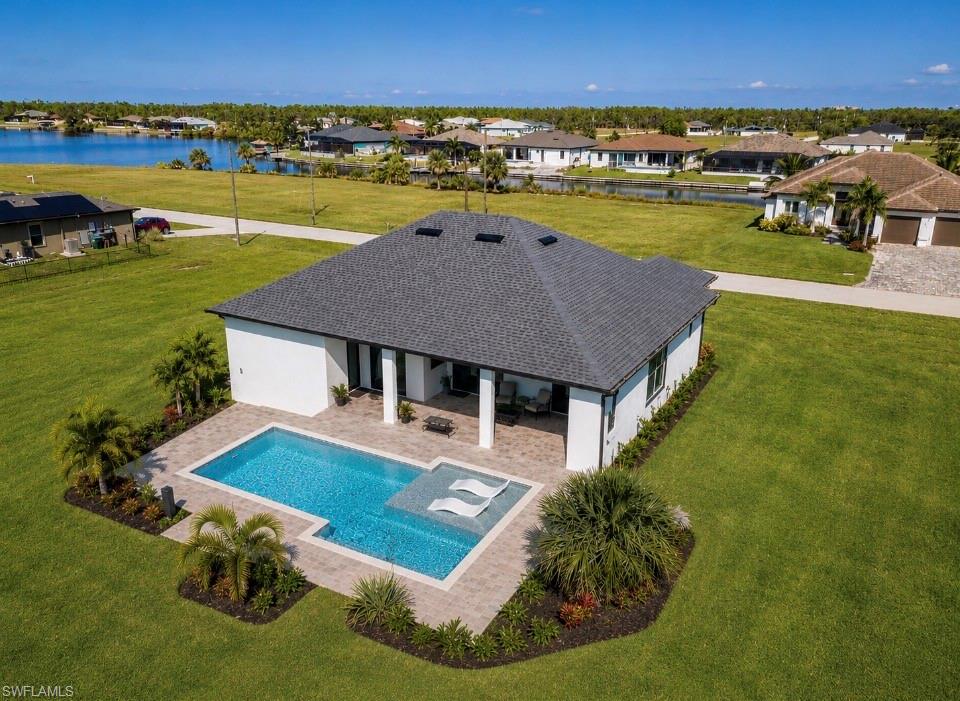 Modern residence with a dark shingle roof and white exterior, featuring a rectangular swimming pool with an integrated sun shelf, extensive paver patio, and established landscape beds