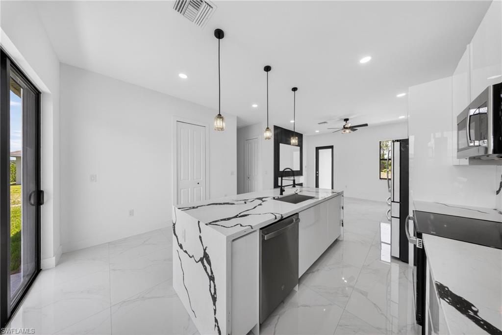Modern kitchen featuring a large island with waterfall-edge countertop, stainless steel appliances, white cabinetry, polished stone flooring, pendant lighting, and an open layout with ceiling fan and recessed lighting throughout