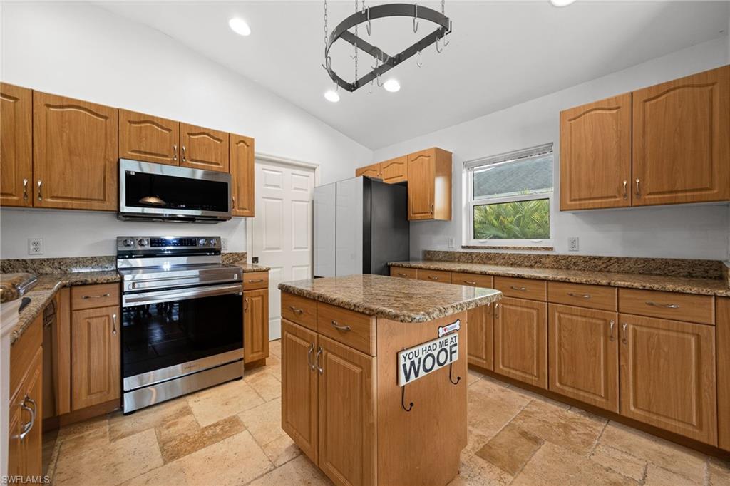 Spacious kitchen featuring warm wood cabinetry, stainless steel appliances including a double wall oven and microwave, granite countertops, a large center island with seating, tile flooring, and abundant natural light from multiple windows
