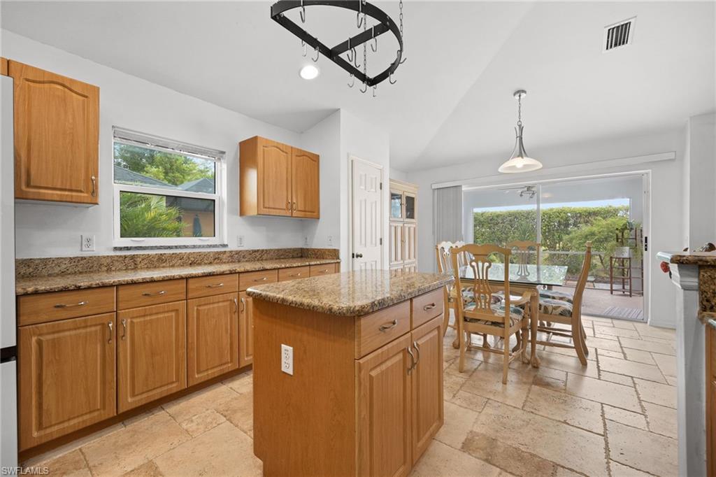Bright kitchen and dining area featuring warm wood cabinetry, granite countertops, a center island with prep space, stainless steel appliances, tile flooring, pendant lighting, and direct access to a covered outdoor patio through French doors