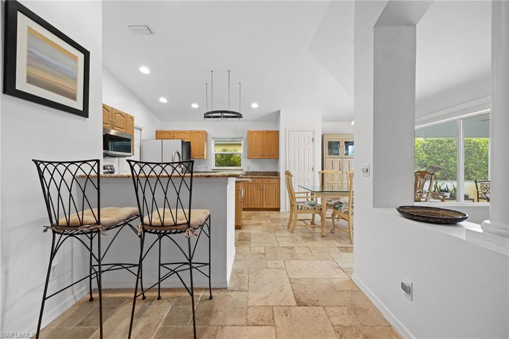 Open-concept kitchen and dining area featuring wood-finish cabinetry, stainless steel appliances, a central island with seating, tile flooring throughout, and abundant natural light from large windows overlooking the outdoor space