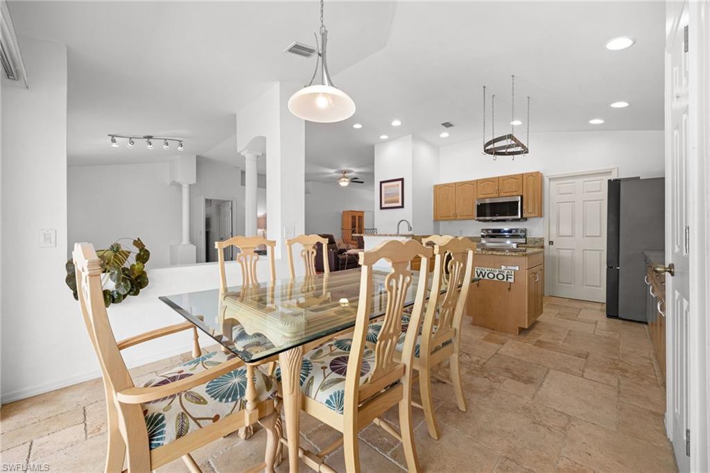 Open-concept dining and kitchen area featuring a glass-top dining table with wood-finish chairs, pendant lighting, stainless steel appliances, wood-finish cabinetry, black refrigerator, and wood-finish flooring throughout