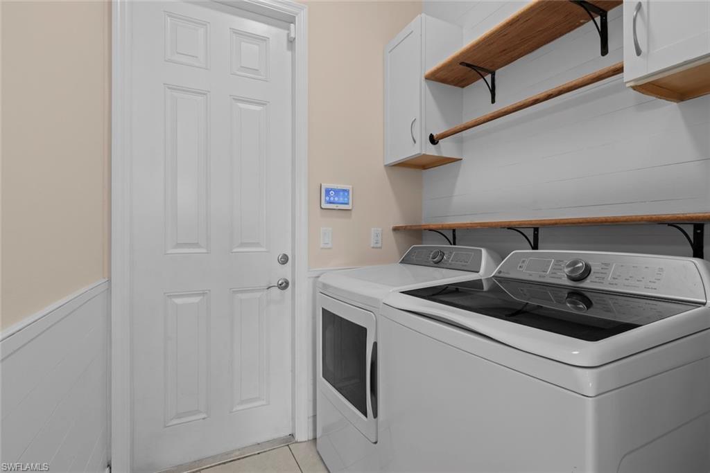 Dedicated laundry room featuring a top-load washer and electric dryer, tile flooring, white cabinetry with wood-finish floating shelves, and garage access for convenient utility management