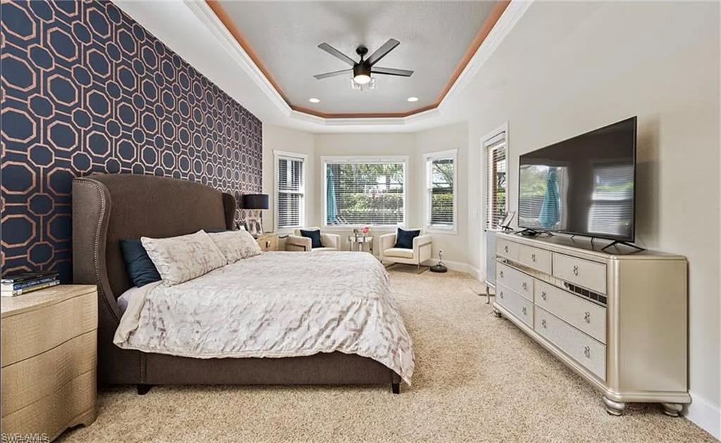Tray ceiling with crown molding, patterned accent wall, bay window, ceiling fan, and carpeted flooring