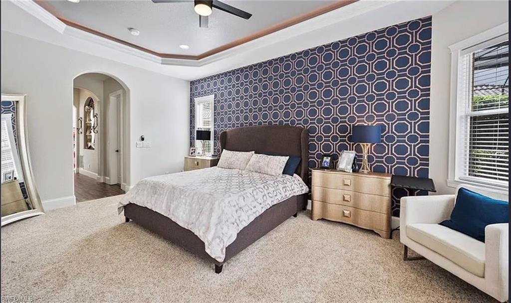 Tray ceiling with crown molding, patterned accent wall, archway entry, and window with blinds