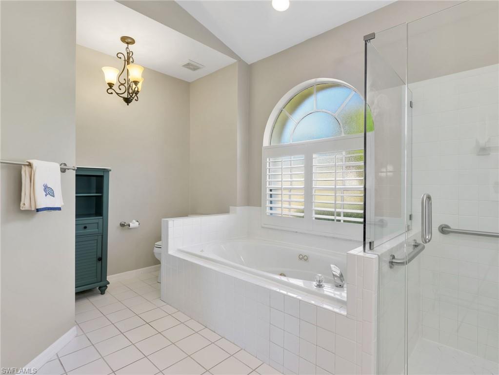 Soaking tub with white tile surround, arched window featuring plantation shutters, glass-enclosed shower, and tile flooring