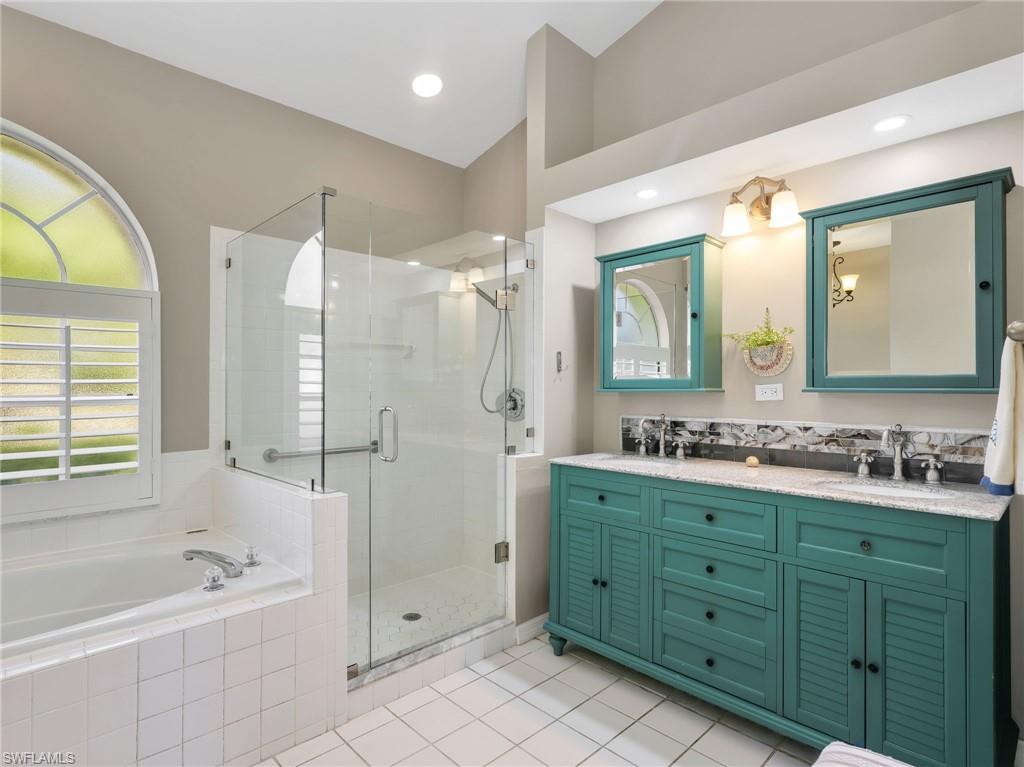 Walk-in shower with glass enclosure, separate soaking tub, double vanity with granite countertop, double medicine cabinets, and arched window with plantation shutters