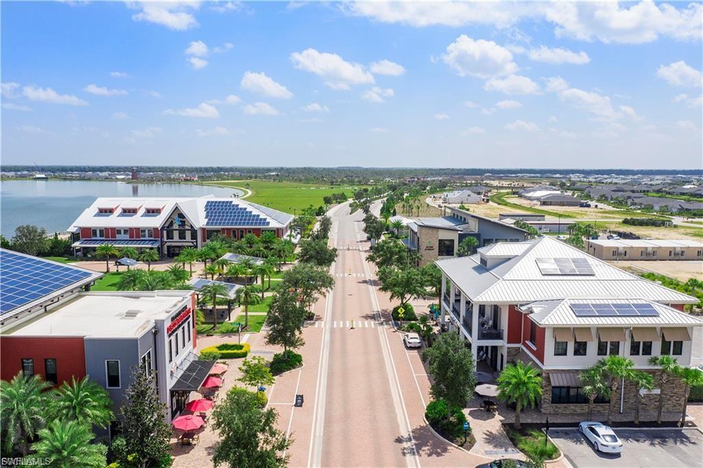 Aerial view showcasing a modern residential community featuring contemporary architecture with mixed red and white exterior finishes, solar panel installations, manicured landscaping with mature trees lining a central boulevard, and expansive green spaces overlooking agricultural countryside and waterfront in the distance