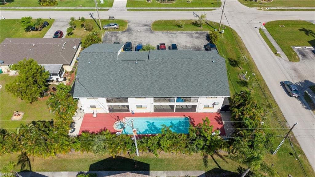 Aerial view of a residential property featuring a spacious single-story home with a charcoal-gray asphalt shingle roof, white exterior siding, and a striking red-finished pool deck with in-ground swimming pool