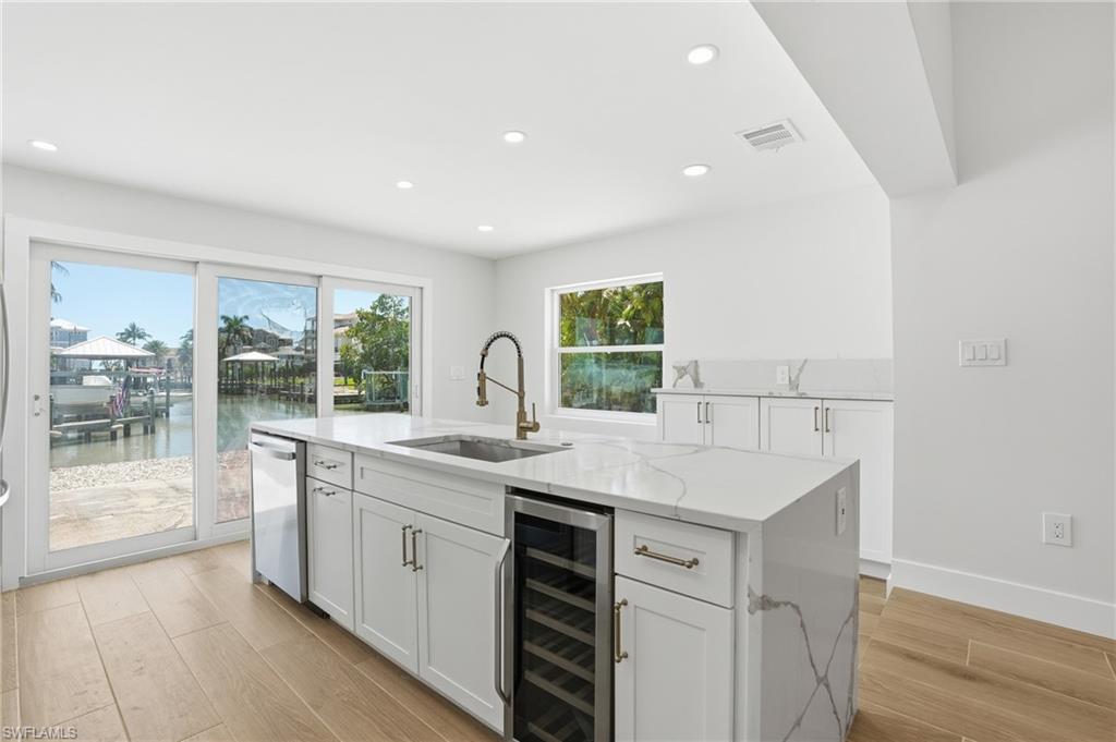 Kitchen featuring an island with a built-in sink, dishwasher, and beverage cooler, white cabinetry, and light-colored countertops - Virtually Edited Image