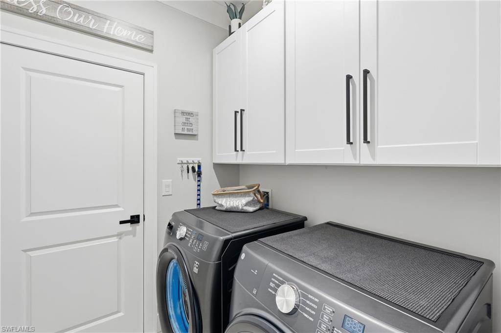 Dedicated laundry room featuring front-load washer and dryer with stainless steel finish, soft-close white cabinetry with brushed metal hardware, quartz countertop work surface, and direct access to the home's interior via solid-panel entry door