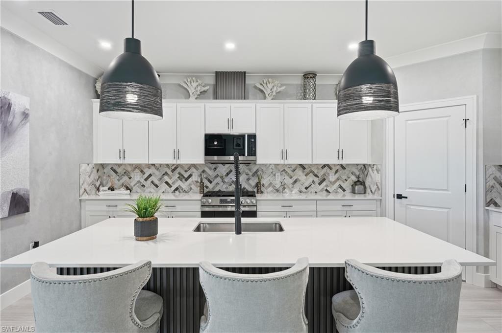 Contemporary kitchen featuring white cabinetry with brushed nickel hardware, a geometric marble tile backsplash, stainless steel appliances including a built-in range, quartz waterfall island countertop with seating for three, and pendant lighting with metal shades suspended above the workspace