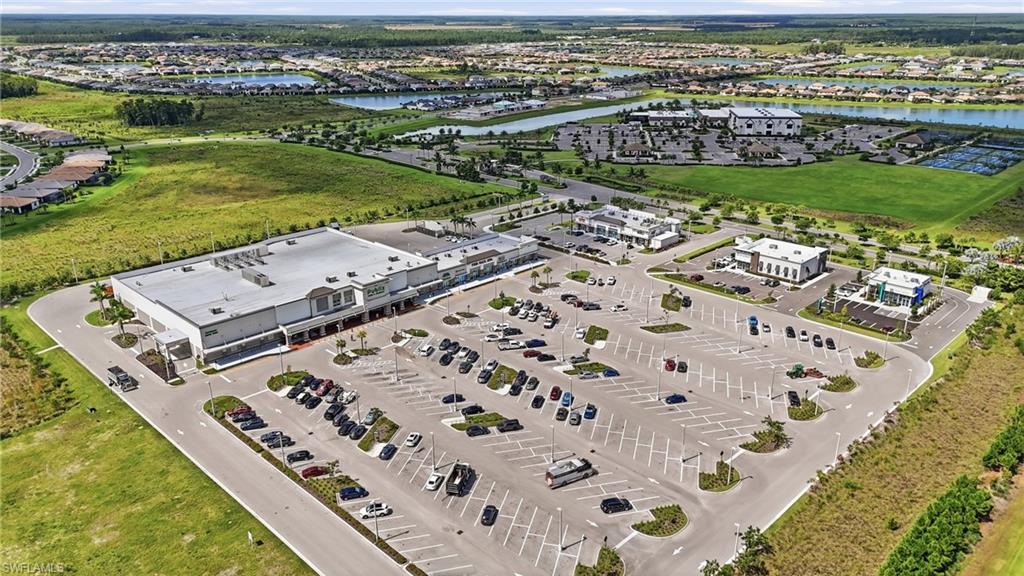 Modern commercial facility featuring an expansive asphalt parking lot with organized striping and landscaped perimeter plantings, a large warehouse structure with a light-colored roof and contemporary architecture, and additional administrative buildings positioned across the manicured grounds