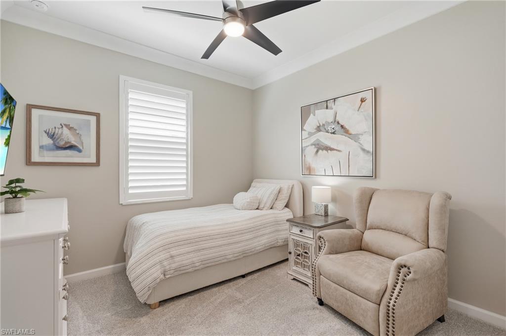Serene bedroom retreat featuring neutral décor, ceiling fan with integrated lighting, plantation shutters on transom window, plush upholstered accent chair with nailhead trim, and soft striped bedding on a platform bed frame