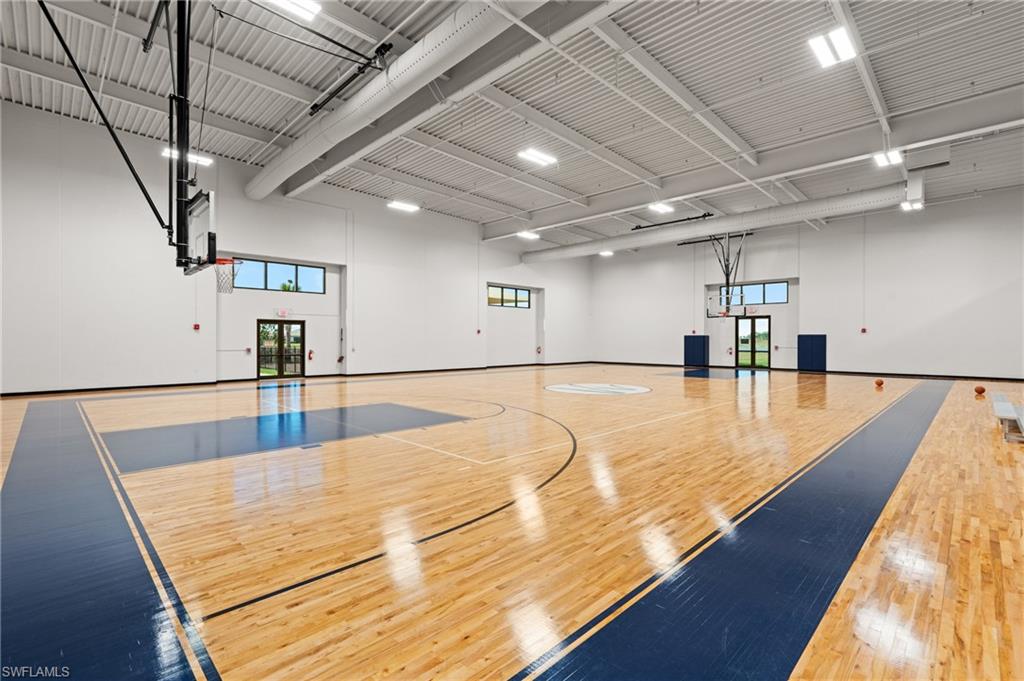 Bright indoor athletic facility featuring a professional-grade wood-finish court with blue lane markings, soaring ceiling with exposed structural beams and recessed lighting, multiple basketball hoops, and large transom windows providing natural illumination throughout the open floor plan