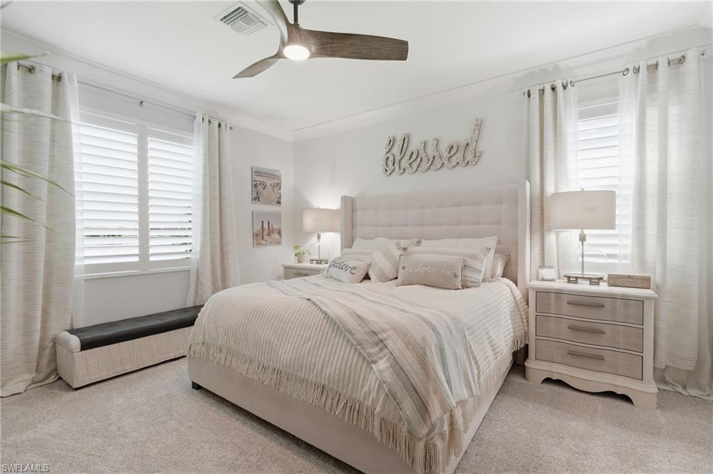 Master bedroom featuring a neutral upholstered headboard with coordinating bedding, ceiling fan with integrated lighting, dual nightstands with attached lamps, plantation shutters on dual windows, and neutral-toned carpet flooring throughout